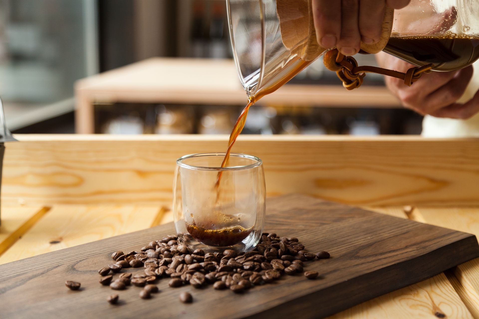 Coffee shop concept represented by barista hands serving the liquid on a crystal glass surrounded by natural coffee beans on a wooden cafeteria environment. 